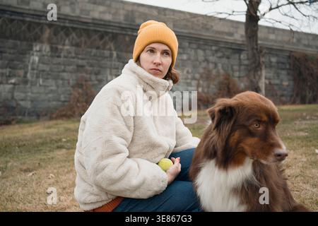 Una donna concentrata in una giacca invernale allena il suo pastore australiano all'aperto, tenendo una palla da tennis durante la pratica dell'obbedienza in un parco, mettendo in risalto il legame Foto Stock
