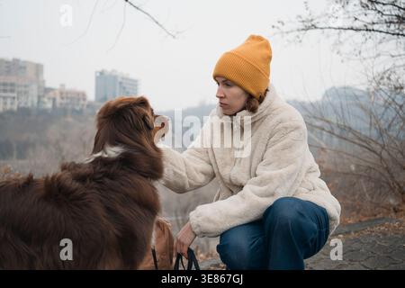 Donna con giacca calda e berretto che cammina un pastore australiano in una giornata piovosa e nebbiosa, fermandosi per confortare il suo cane su un sentiero bagnato con un paesaggio urbano Foto Stock