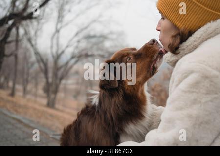 Primo piano di una donna con un cappotto caldo e un berretto che abbraccia il suo pastore australiano all'aperto in inverno, catturando la lingua del cane mentre le lecca il viso in un Foto Stock