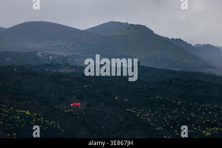 Veduta in modo Moody del paesaggio vulcanico di Cumbre Vieja a la Palma, Isole Canarie, con campi di lava scura e un'auto rossa solitaria su una strada sotto i monti nebbiosi Foto Stock