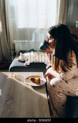Accogliente mattina al coperto a casa, donna in pigiama sorseggiando un caffè vicino a una finestra illuminata dal sole, mentre un gatto bianco dorme sul divano, con torta sul tavolo, calma e. Foto Stock
