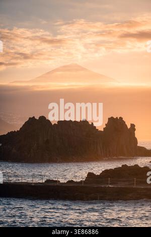 Calda vista del tramonto da El Pris sulla costa settentrionale di Tenerife, con il monte Teide che si innalza sopra la nebbia del mare. Spettacolari rocce vulcaniche, calme acque atlantiche e sof Foto Stock