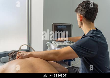 Il fisioterapista esegue l'ecografia diagnostica sulla schiena di un paziente in una clinica moderna, toccando il pannello di controllo e osservando il monitor mentre guidi Foto Stock
