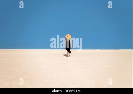 Una donna con un abito nero e un cappello di paglia cammina attraverso una vasta duna di sabbia sotto un cielo azzurro e limpido a Corralejo, Fuerteventura, parte della bellissima lattina spagnola Foto Stock