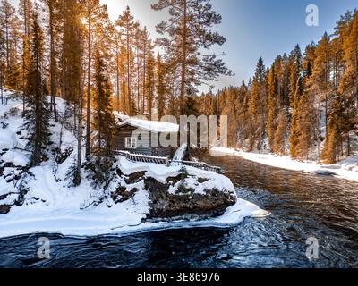 Accogliente cabina in legno su una sponda innevata circondata da alti pini, dalla calda luce dell'alba e dal cielo invernale limpido in Finlandia, catturando tranquille zone selvagge Foto Stock