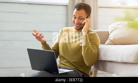 Uomo che parla al telefono seduto a terra con un laptop in uno spazio abitativo moderno Foto Stock