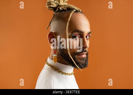 Uomo gay nero sicuro di sé con trucco luminoso in piedi isolato su sfondo arancione, vestito di bianco. Trasuda senso di orgoglio e individualità. Potere di diversità dello stile personale. Primo piano Foto Stock