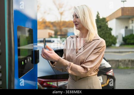 L'utente interagisce con la stazione di ricarica del veicolo elettrico in ambiente urbano. Una moderna auto nera è parcheggiata nelle vicinanze ed è visibile un cielo azzurro. Foto Stock