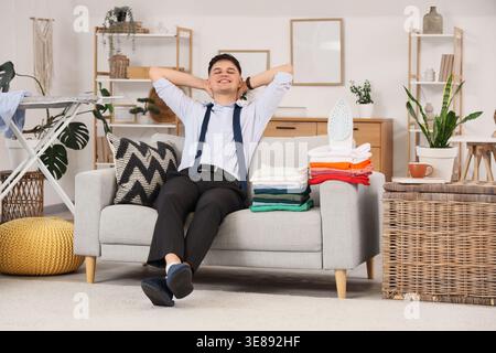 Bel giovane uomo d'affari felice con ferro da stiro e pile di vestiti seduti sul divano di casa Foto Stock
