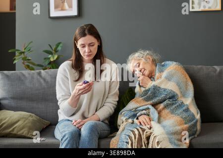Donna adulta caucasica che controlla il termometro mentre la donna anziana riposa sotto la coperta e usa l'inalatore, mostrando cure di malattia a casa. Utile per annunci sanitari e campagne di assistenza sanitaria Foto Stock
