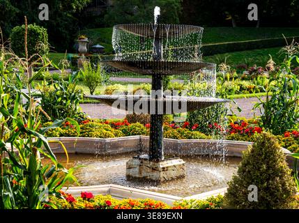 Fontana in pietra a due piani che si innalza in un giardino ben curato con aiuole colorate, vegetazione e un parco soleggiato. Foto Stock
