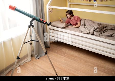 Ragazza sdraiata a letto con smartphone con telescopio in piedi nelle vicinanze. Foto Stock