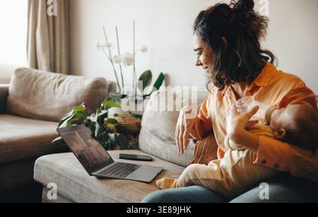 Una mamma si siede su un comodo divano a casa, dando da mangiare al suo bambino mentre guarda un notebook e sorride. Un momento familiare caldo e rilassato con piante nelle vicinanze. Foto Stock