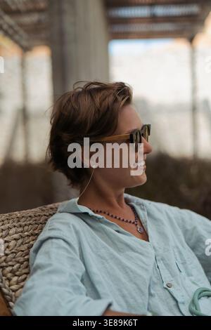 Donna felice con gli occhiali da sole sorridente di profilo seduta all'aperto su una sedia Foto Stock