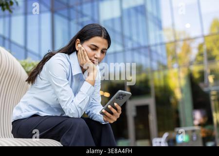 Giovane donna d'affari con un auricolare seduto all'aperto su una panchina moderna, che guarda il suo smartphone con un'espressione noiosa mentre aspetta qualcuno o qualcosa vicino a un edificio aziendale in vetro Foto Stock