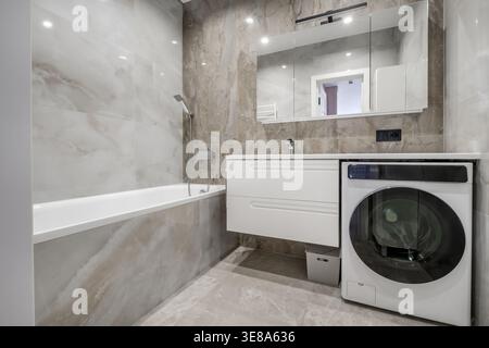 Interni luminosi e minimalisti del bagno con lavello in ceramica bianca, vasca da bagno, rubinetto a parete e corrimano di sicurezza sulla parete piastrellata. Foto Stock