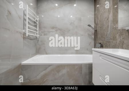 Interni luminosi e minimalisti del bagno con lavello in ceramica bianca, vasca da bagno, rubinetto a parete e corrimano di sicurezza sulla parete piastrellata. Foto Stock