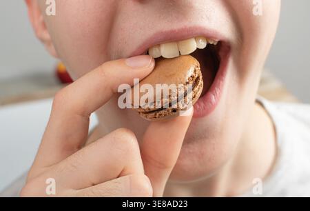 Un bambino mette in bocca una torta di Macaron sorridente, biscotto di Macaron in mano primo piano, meringa di mandorle, macaron dolci, dessert francese colorato Foto Stock