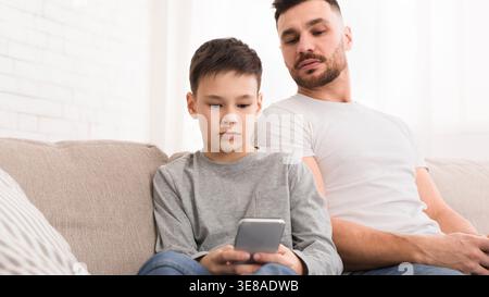 Padre siede con figlio su un divano mentre il bambino guarda lo schermo di uno smartphone Foto Stock