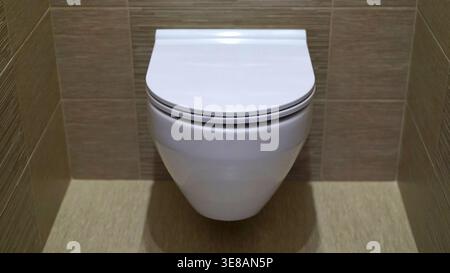 WC moderno a parete con coperchio chiuso, centrato in un bagno minimalista. La ceramica bianca pulita contrasta con le pareti piastrellate di marrone beige neutro. Symmetri Foto Stock