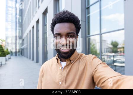 Il giovane imprenditore nero sorridente scatta un selfie sicuro all'aperto in un ambiente urbano moderno, con la testa candida con gli edifici degli uffici sullo sfondo luminoso diurno, autentico e allegro Foto Stock