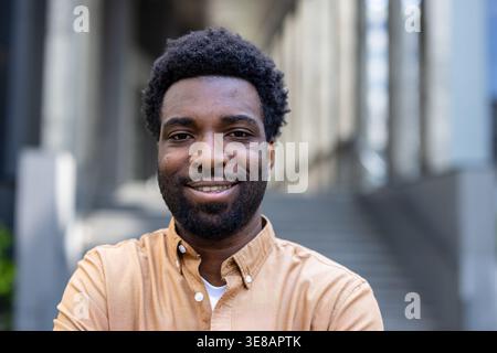 Giovane sorridente alla telecamera con un'espressione autentica, che trasmette positività e successo mentre si trova in un ambiente urbano contemporaneo con dettagli architettonici sullo sfondo Foto Stock