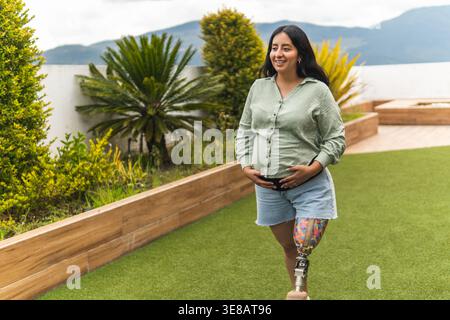 Donna latinx incinta con una gamba protesica che cammina all'aperto, tenendo il bambino a batti e sorridendo Foto Stock