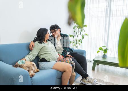 Donna incinta con gamba protesica e partner che si rilassa su un divano a casa con il proprio cane Foto Stock