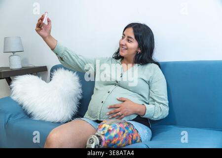 Donna incinta con una gamba protesica seduta su un divano che fa un selfie a casa Foto Stock