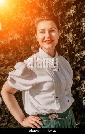 Donna sorridere in giardino donna adulta sorridente in camicetta ricamata che gode della luce del sole dorata in un lussureggiante giardino verde Foto Stock