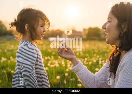 Madre e figlia soffiano il dente di leone durante una passeggiata all'aperto nella natura, sorridendo e legando, concetto di maternità e giorno della mamma Foto Stock