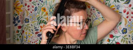 Una donna di 50 anni si trova in un bagno con una tenda doccia floreale. Utilizza una piastra per capelli, concentrandosi sui capelli mentre si prepara per il Foto Stock