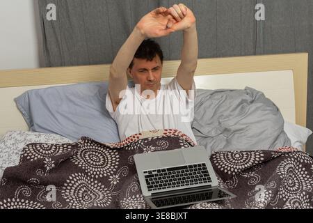 Un uomo si allunga mentre si siede a letto con un portatile davanti a sé. È mattina e si è appena svegliato. Il letto sembra disordinato con coperte e lenzuola intorno a lui. Foto Stock