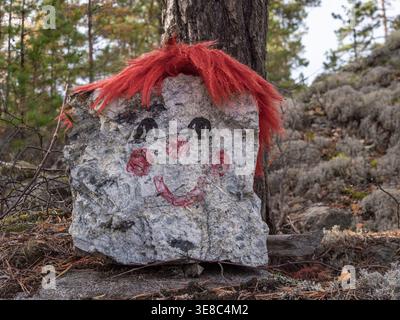 Roccia dipinta con una faccia sorridente e parrucca rossa appoggiata contro un albero in una radura della foresta Foto Stock