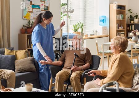 Uomo caucasico anziano che riceve un trattamento IV da infermiera in casa di cura, seduto con la canna e interagendo con il gruppo. Utile per assistenza medica, assistenza sanitaria domiciliare, marketing della vita assistita Foto Stock