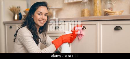 Una giovane donna pulisce la cucina indossando guanti di gomma e sorridendo in un ambiente Bright Home Foto Stock