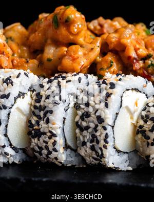 I rotoli ricoperti di sesamo si trovano accanto a un piatto di pollo piccante. Il riso ricco e cremoso contrasta con texture audaci e croccanti. La piastra scura evidenzia colori vivaci e. Foto Stock