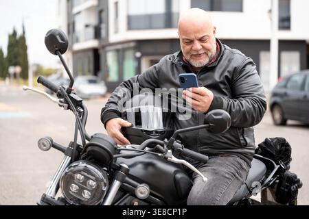 Uomo anziano che utilizza un telefono cellulare, un social media o un'app di navigazione, riposa sulla sua moto in un ambiente urbano Foto Stock