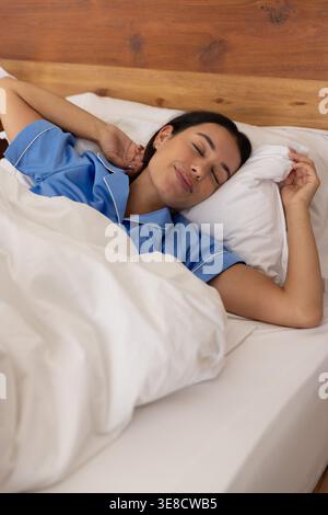 Donna sdraiata in camera da letto sotto trapunta bianca che regge il cuscino contro la testiera indossando pigiami blu Foto Stock