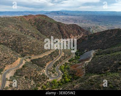 Vista aerea di una spettacolare strada tortuosa che serpeggia tra le colline di roccia rossa e la vegetazione verde vicino a Jerome, Arizona, sotto un cielo parzialmente nuvoloso. Foto Stock