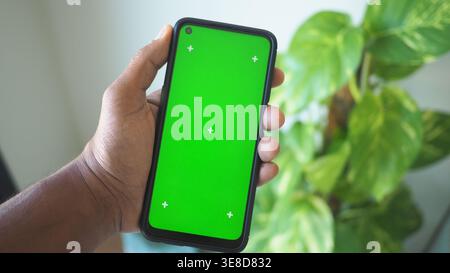 Mano che tiene lo smartphone con schermo verde in ambiente interno Foto Stock
