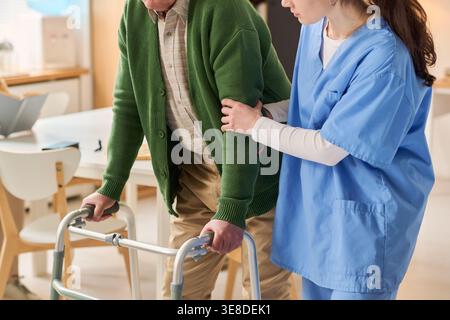 Uomo anziano che utilizza un deambulatore mentre una giovane donna adulta assiste alla mobilità durante la riabilitazione. Ideale per assistenza agli anziani, assistenza sanitaria, supporto al recupero Foto Stock