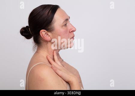 Una donna sta di profilo, toccando delicatamente il collo con un'espressione premurosa. Il semplice sfondo migliora il suo comportamento calmo, suggerendo un momento Foto Stock