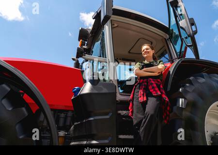 Donna agricoltrice in piedi con le braccia incrociate vicino al trattore all'esterno, vista ad angolo basso. Foto Stock
