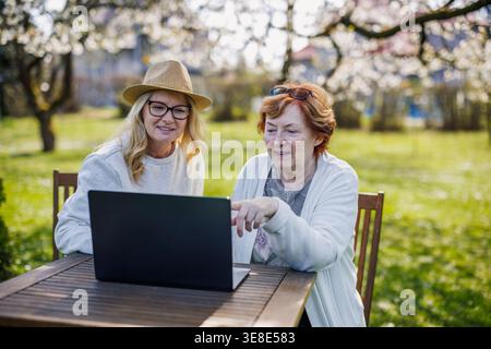 Figlia adulta che mostra il laptop a una madre anziana nel giardino di primavera. Una famiglia plurigenerazione felice che utilizza la tecnologia insieme durante le giornate soleggiate all'aperto Foto Stock