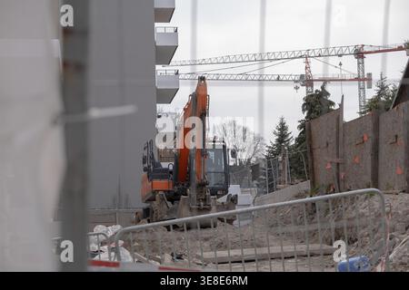Escavatore operativo in cantiere urbano con gru e edifici residenziali in background. Concentrarsi sullo sviluppo attivo, sui macchinari pesanti e b Foto Stock