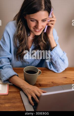 Giovane donna che lavora in multitasking con smartphone, notebook, caffè e appunti in casa. Lavoro remoto, produttività, comunicazione digitale e freelan moderno Foto Stock
