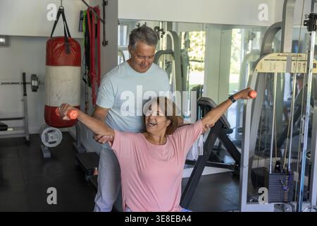 Donna anziana in cima rosa che solleva manubri arancioni sulla panchina mentre uomo sostiene braccia, specchi da palestra Foto Stock