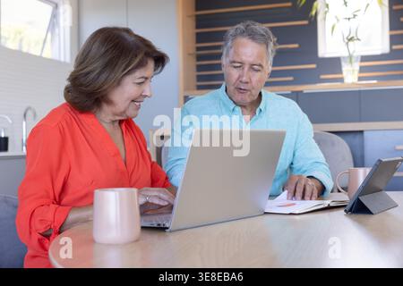 Donna anziana in camicetta di corallo che scrive su un notebook argento mentre l'uomo controlla il notebook al tavolo della cucina Foto Stock