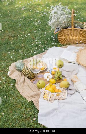 Primo piano di un cesto di vimini con un mucchio di fiori di respiro, un libro aperto, cappello di paglia, baguette fresche, formaggio, ananas, frutta assortita e bicchieri da vino su una coperta da picnic in un giardino estivo Foto Stock